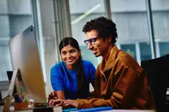 Male with blue glasses and female looking at a computer screen sitting at a desk