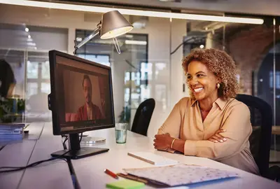 Female smiling, sitting at a desk behind a computer screen in a video meeting