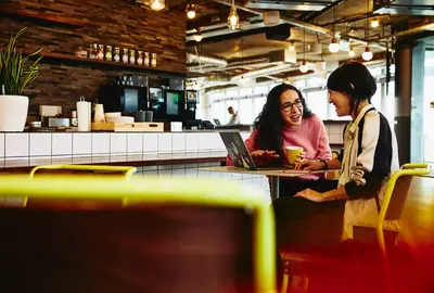 Two female sitting in restaurant having a converstation, looking at laptop, smiling.
