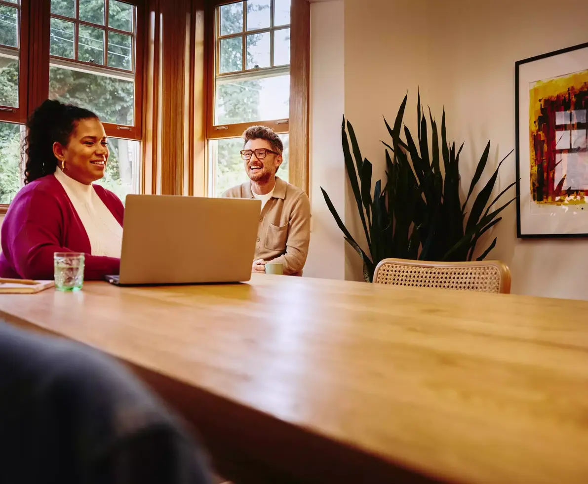 Female sitting at a table looking at her laptop with smiling male next to her at home
